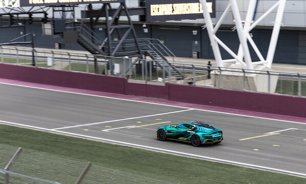 Sports car driving past the pit lane at Silverstone Circuit, with crews and support vehicles in the background.