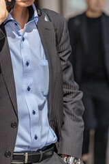Mobile phone to ear, stylish businessman in gray pinstripe suit and light blue checkered shirt conducts business while walking among people in urban setting.
