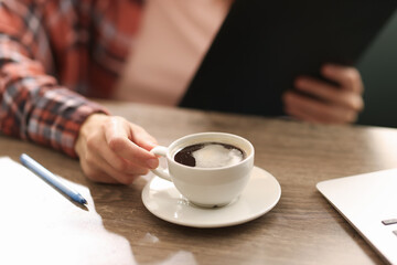 Coffee break. Woman with cup of aromatic drink and clipboard at wooden desk indoors, closeup