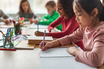 Korean schoolgirl focuses on writing notes during class, sitting with multicultural classmates. The classroom is bright, promoting a modern educational environment full of creativity.