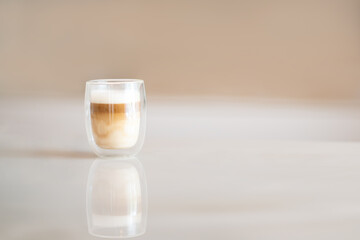 Cappuccino macchiato with creamy milk layer in modern double wall glass cup with reflection on smooth white table