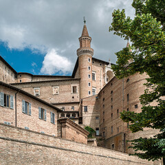 The majestic Palazzo Ducale in Urbino, Marche, Italy, seen from below, is rising from the other buildings. Renaissance brick architecture with a tall tower against a cloudy sky.