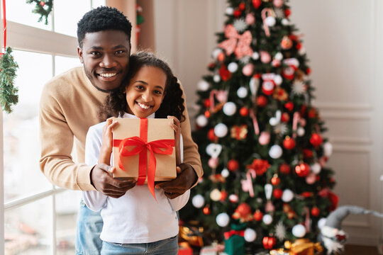 A loving African American family celebrates Christmas together by a festive tree. The father embraces his adorable daughter, who is excitedly holding a gift box and smiling for the camera.