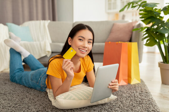 A positive asian woman relaxes on the carpet in her living room. She is using a digital tablet and smiling at the camera while holding a credit card. Brightly colored shopping bags are nearby.
