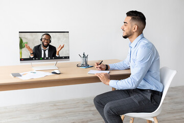 A smiling young Arab man is sitting at a wooden desk with a laptop and notebook, engaging in a video call with a colleague during remote work from a bright office.