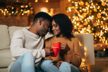 A beautiful young black couple touches foreheads while holding red mugs of coffee. They enjoy a romantic evening together on Christmas Eve in a warmly decorated room filled with festive lights.