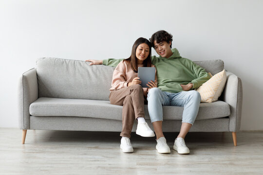 A happy Asian couple sits closely on a gray couch at home, smiling as they engage with a digital tablet. They are exploring a new application together, creating a cozy atmosphere. - Powered by Adobe