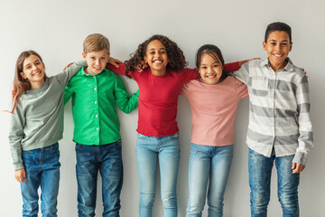 Obraz premium Five cheerful children of different backgrounds are hugging and smiling together indoors. They stand against a gray wall, showcasing the joy of friendship in a playful and lively atmosphere.