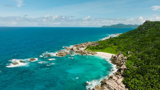 Granite rocks and lush greenery along a turquoise coastline with waves crashing against the shore. Anse Marron beach. La Digue, Seychelles.