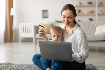 A woman sits on a rug with her young child, using a laptop for online shopping. She holds a credit card, smiling as they enjoy their time together at home.