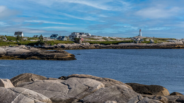 Rocky Atlantic coastline at Cape Breton, Nova Scotia, with blue ocean, seaside houses, and a distant white lighthouse under a bright sky. Peaceful coastal landscape.