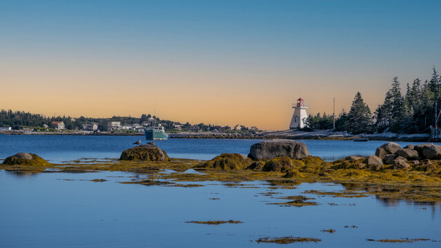 Calm coastal inlet at Cape Breton, Nova Scotia, with low tide rocks, a moored fishing boat, and a white lighthouse at sunset under a clear sky.