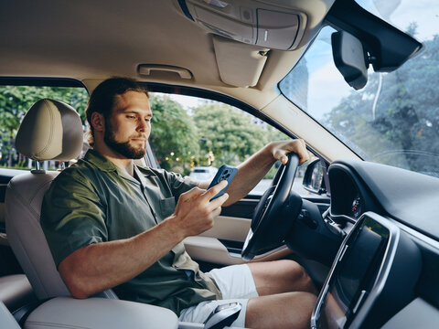 A focused man sits in a car, glancing at a smartphone while driving. Casual attire, modern interior, clean dashboard, and daylight create a calm, everyday moment.