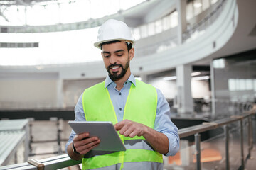A construction worker in a bright yellow safety vest and helmet stands near a railing indoors, attentively using a tablet to review plans or monitor progress at the building site.