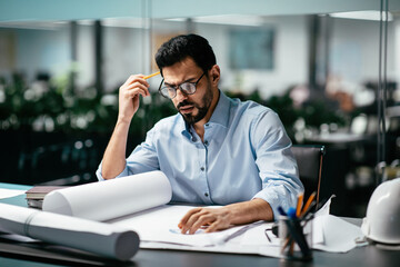 A focused architect sits at a desk covered with rolled blueprints and sketching materials. He...
