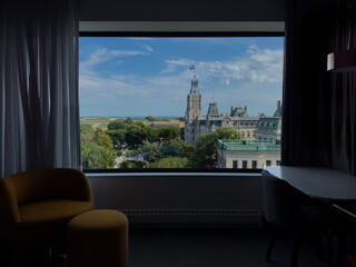 View of the Quebec Parliament Building and nearby historic district framed by a hotel window, with blue sky and greenery outside and modern interior elements in silhouette.