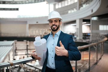 A man in a hard hat and suit smiles and shows a thumbs up while holding architectural blueprints in...