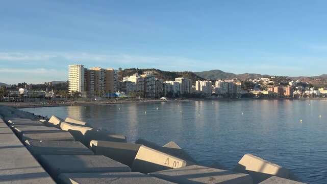 Concrete wave breakers along promenade with calm sea, distant lighthouse, coastal buildings, and mountain backdrop creating relaxed Mediterranean shoreline atmosphere during daytime