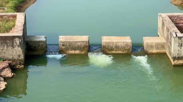 A close-up view of a small concrete check dam holding calm green water with gentle flow through narrow gates, surrounded by rocks and greenery, showcasing rural irrigation and water management.