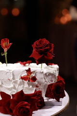 Fototapeta premium Close up of rectangular white wedding cake with textured icing, red roses and unlit candles on wooden table, dark wall background. Concept of wedding, romance, luxury, celebration.