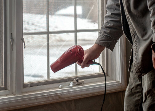  A hand holds a blow dryer and is being used to Shrink wrap a window, to help insulate and keep in the heat for winter