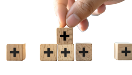 Hand placing a plus block atop a small wooden tower. Other blocks with plus signs are nearby