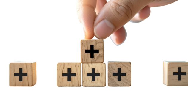Hand placing a plus block atop a small wooden tower. Other blocks with plus signs are nearby