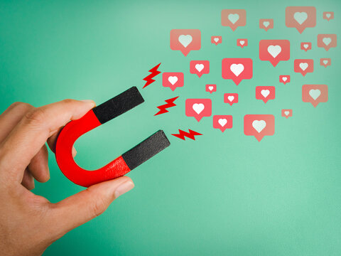 Hand holding red magnet attracting social media like icons, symbolizing digital marketing, content engagement, social influence, and online audience connection on green background. - Powered by Adobe