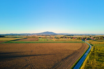 Obraz premium Aerial landscape with green and brown agricultural fields, road, small village and large mountain under clear blue sky