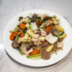 Stir-fried noodles topped with mixed vegetables, chicken, and meatballs, served on a white plate. Colorful and appetizing Asian cuisine.