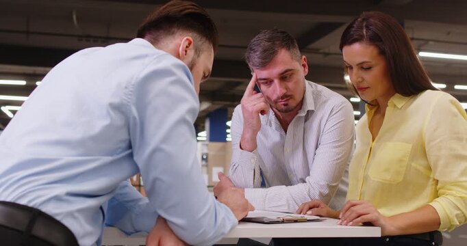 Professional male salesman and young couple standing in office and discussing terms of contract. Focused clients talking to agent, exploring options and reviewing document before signing agreement.