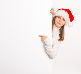 Cheerful preteen girl in Santa hat smiling and peeking from behind white wall while pointing at copy space. Christmas holiday concept isolated on white background.