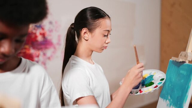 Multicultural student paint the canvas together in art lesson with colorful stained wall. Closeup of african boy and caucasian girl drawing picture or doing creative activity together. Edification