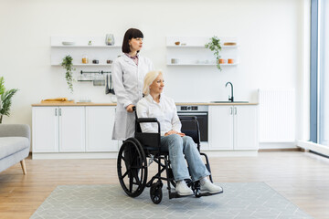 A female caregiver pushes an elderly woman in a wheelchair through a modern kitchen, providing care...