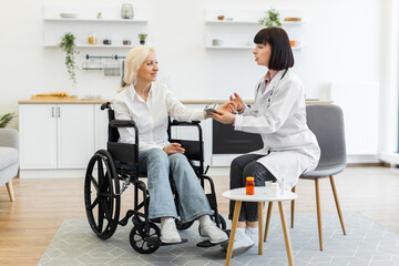 A female doctor examines an elderly woman's blood pressure while she sits in a wheelchair at home, discussing her health.