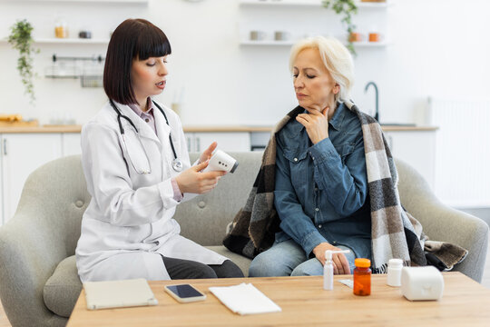 A female doctor is checking an elderly patient's temperature using a digital thermometer while the patient sits on a couch.