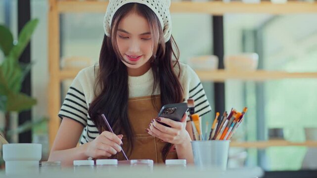 Cheerful Asian female artist using smartphone to find references and ideas for painting ceramic pottery design in creative studio, woman painting custom artwork for client project