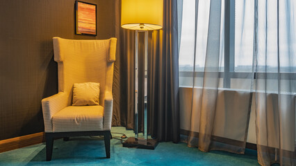 Details of the living room interior. A soft armchair in the corner. A cushion on the seat. A lighted floor lamp nearby. Blue carpet on the floor. Translucent tulle curtains on the window.