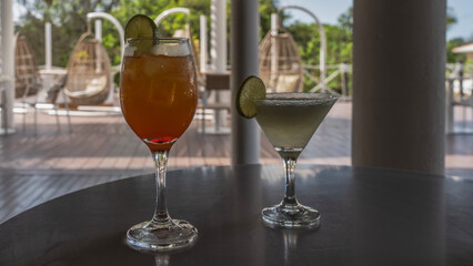 Two glasses with cold cocktails on the table in an outdoor cafe. Orange and green drinks with ice cubes and lime wedges on the edge. The soft background- a terrace, hanging swing chairs, vegetation  