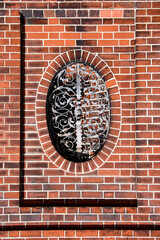 An architectural close-up showing the detail of a classic red brick wall with an oval window or decorative cutout protected by an ornate wrought iron grille. Traditional german construction.