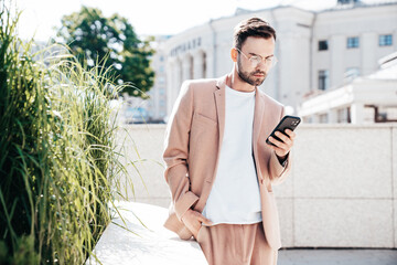Handsome smiling stylish lambersexual model. Sexy modern man dressed in elegant beige suit. Fashion male posing in the street Holds mobile phone, using smartphone apps, looks at telephone screen © halayalex