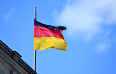 The German flag with black, red, and gold stripes flying at half-mast on a flagpole against a bright blue sky with white clouds and the corner of a building. A symbol of mourning or remembrance.