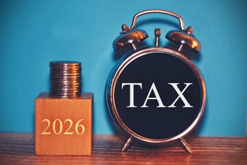 Wooden block with stack of coins symbol of Taxes 2026 text with alarm clock on blue background