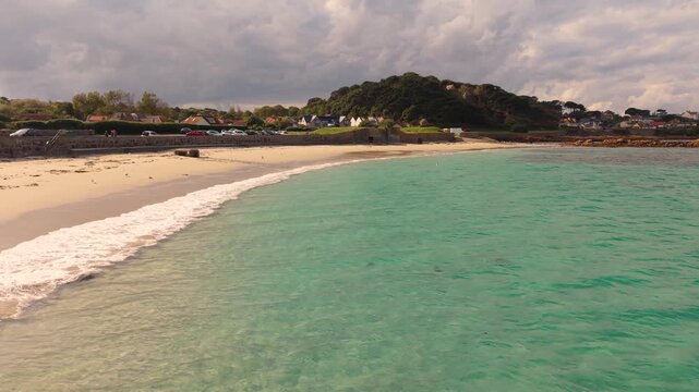Crystal clear water and golden beach at the stunning Cobo Bay Guernsey Channel Islands with clear turquoise water and lapping waves in the sunshine with dramatic sky
