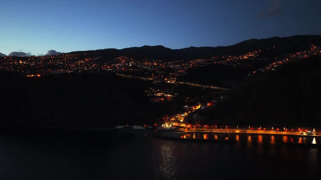 Illuminated Calheta town and harbor at twilight, city lights dot hillside by calm sea, Madeira, Portugal. Aerial forward,copy space