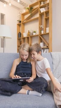 Girl plays game on tablet while boy sits beside helping and cheering, both smiling and celebrating victory together