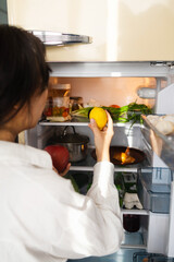 Back view of brunette woman standing in front of opened refrigerator full of food products, choosing ingredients to cook nutritious healthy breakfast before going to work