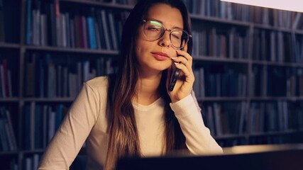 Side view of a student taking a phone call during study time at a library table, multitasking, communication and campus life themes. - Powered by Adobe