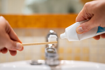 Person's hands holding an ecological bamboo toothbrush and a tube of toothpaste, preparing for daily dental care and sustainable teeth cleaning in a bathroom
