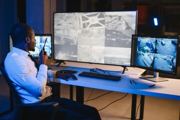 African American security guard looking at desktop computer screen while speaking on walkie talkie